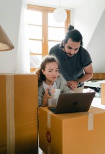 Thoughtful woman surfing internet on netbook while sitting with cup of hot drink near ethnic boyfriend in watch and casual wear standing near cardboard boxes after moving into new flat